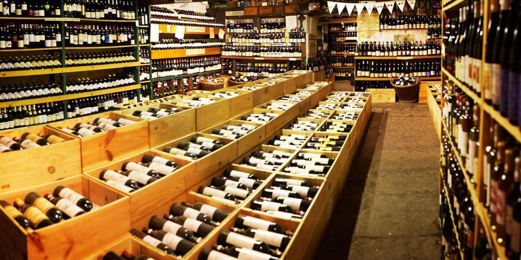 A wine cellar with wooden crates filled with bottles, shelves stocked with wine bottles, and a sign indicating a food section.