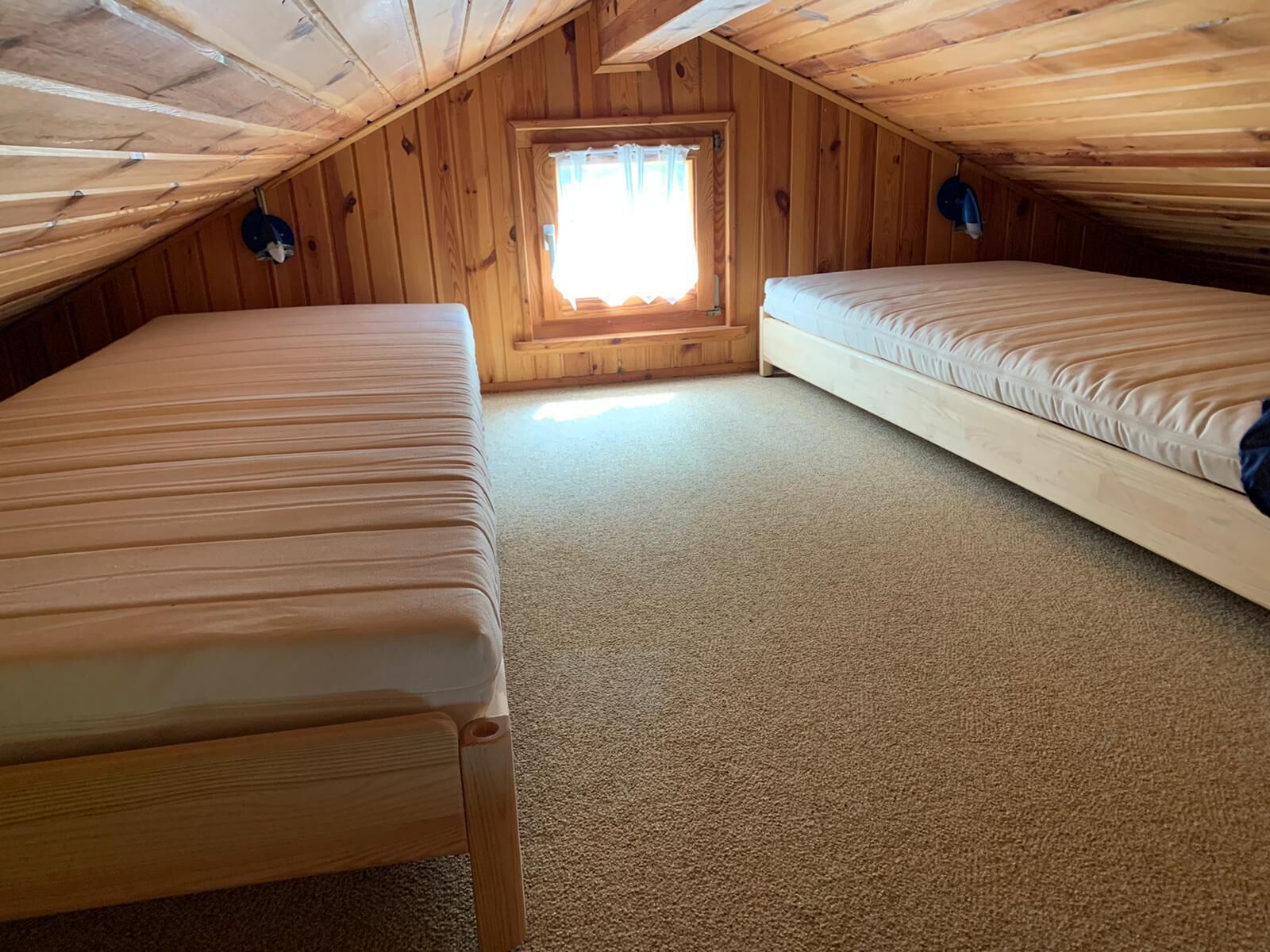 Um pequeno quarto de sótão, único, com duas camas, uma janela com cortinas e chão coberto por carpete.