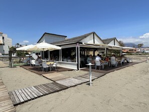 An outdoor café with white tables and chairs, shaded by umbrellas, on a sandy beach with a wooden deck.