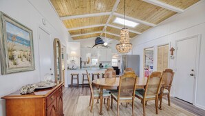 A dining area with a wooden table and chairs, a chandelier, and a kitchen in the background.