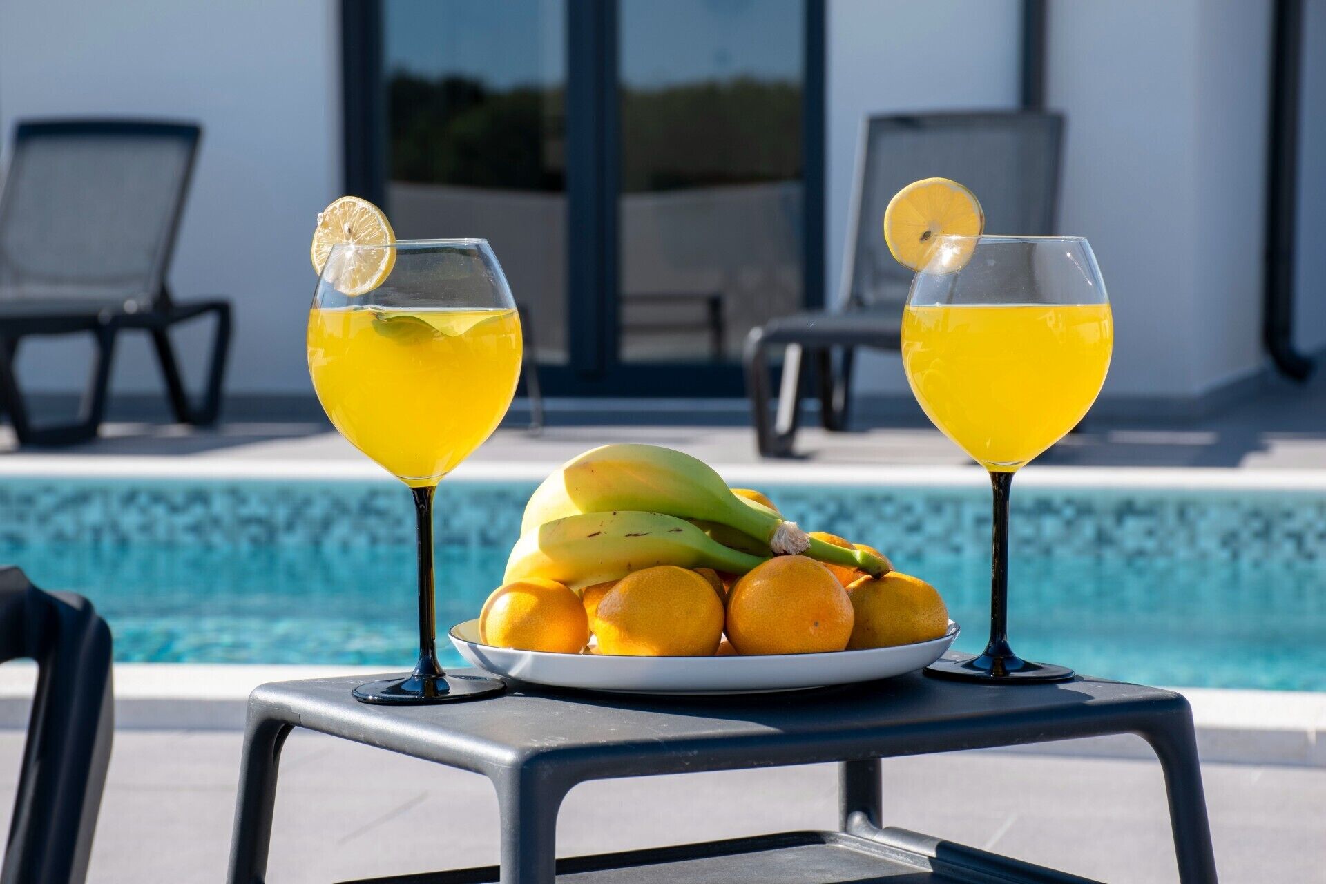 Två glas apelsinjuice med citronskivor står på ett bord vid poolkanten, och en tallrik med apelsiner och bananer står i närheten.