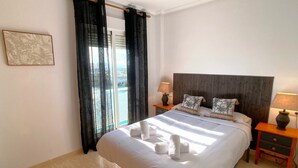 Una cama bien hecha con ropa blanca, dos mesitas de noche con lámparas, un ventanal con cortinas y un cuadro en la pared.