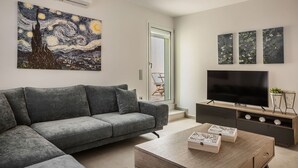 Un soggiorno moderno con un divano grigio, un tavolino in legno e una TV a schermo piatto. Tre opere d'arte incorniciate adornano la parete e un condizionatore d'aria è posizionato sopra il divano.
