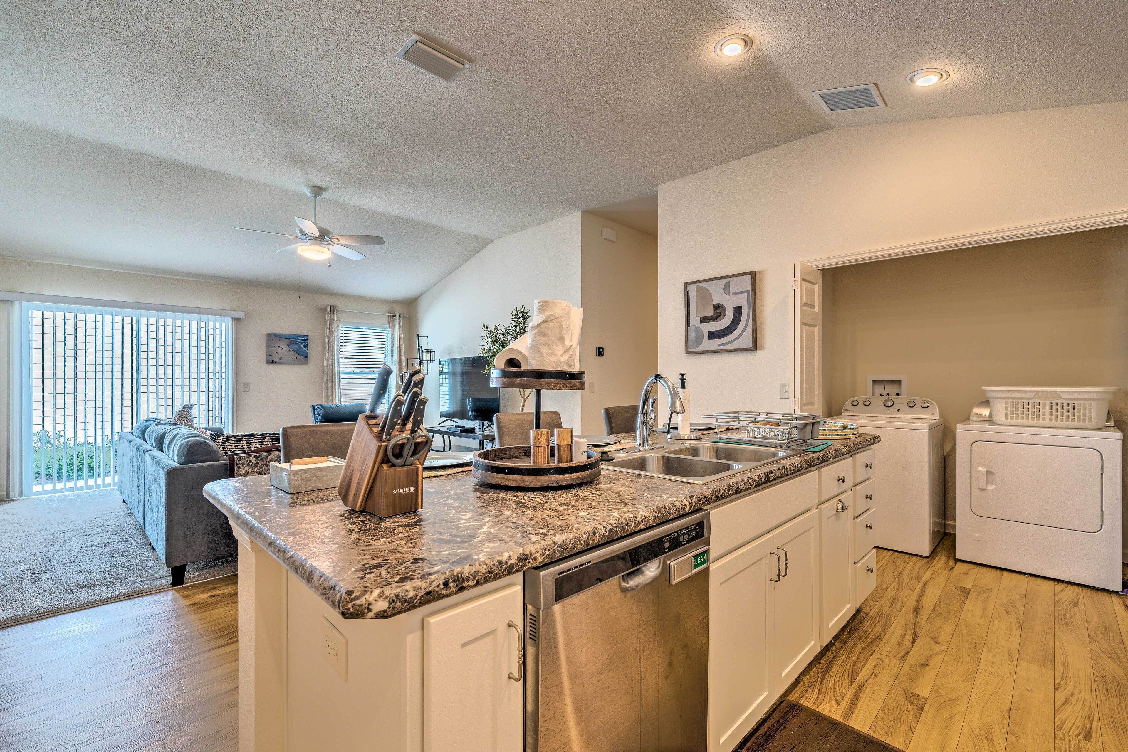 A modern kitchen with granite countertops, stainless steel appliances, and a dishwasher. There is a dining area with a gray sofa and a dining table.