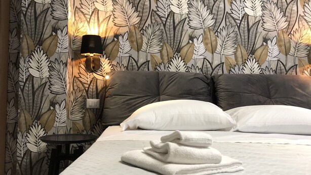 A neatly made bed with white linens and a folded towel, a dark grey upholstered headboard, and a wall with a leafy pattern wallpaper. - Dante 3 Guesthouse (Bari)