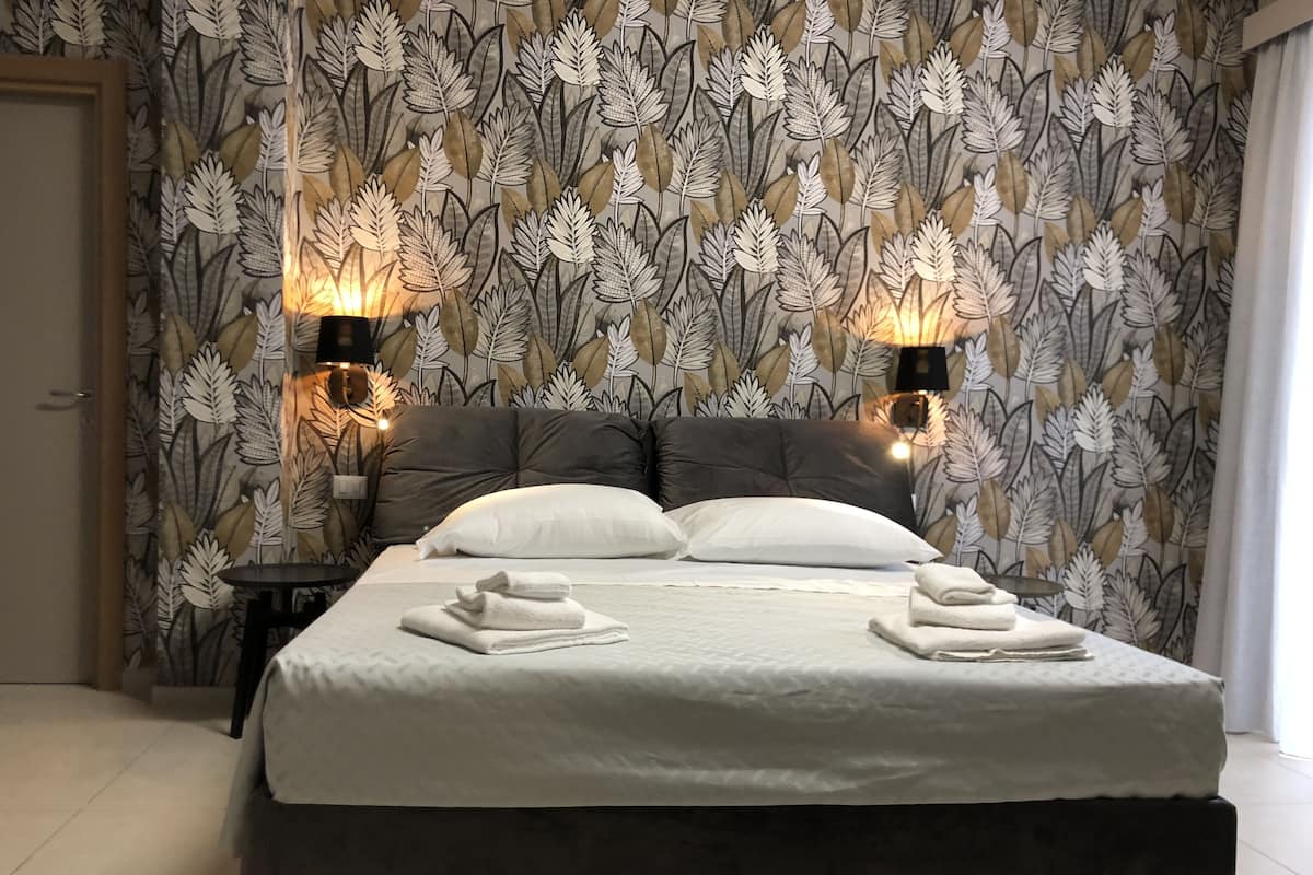 Een net opgemaakt bed met witte lakens en twee kussens, een donkere hoofdbord en een muur met een bladerenpatroon.