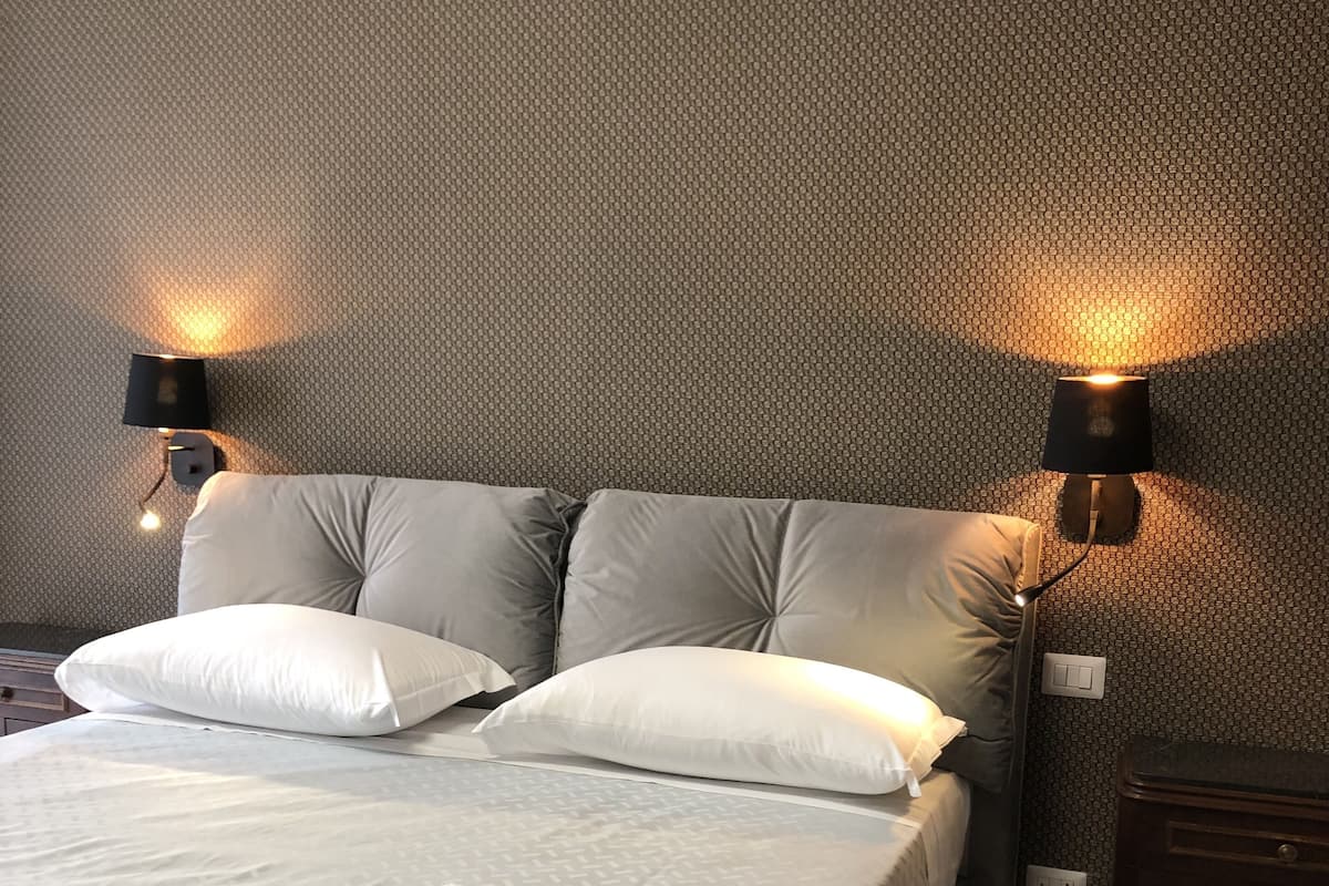 Een net opgemaakt bed met een beddekleed met patroon, twee witte kussens en opgerolde handdoeken op het bed. Aan weerszijden van het bed hangen twee wandlampen.