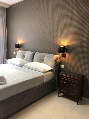 A neatly made bed with a grey duvet and white pillows, two bedside lamps, and a small wooden nightstand with a drawer.