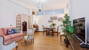 Ein Wohnzimmer mit einem rosafarbenen Sofa, gemusterten Sesseln, einem Esstisch mit Stühlen, einem Fernseher und Holzboden.