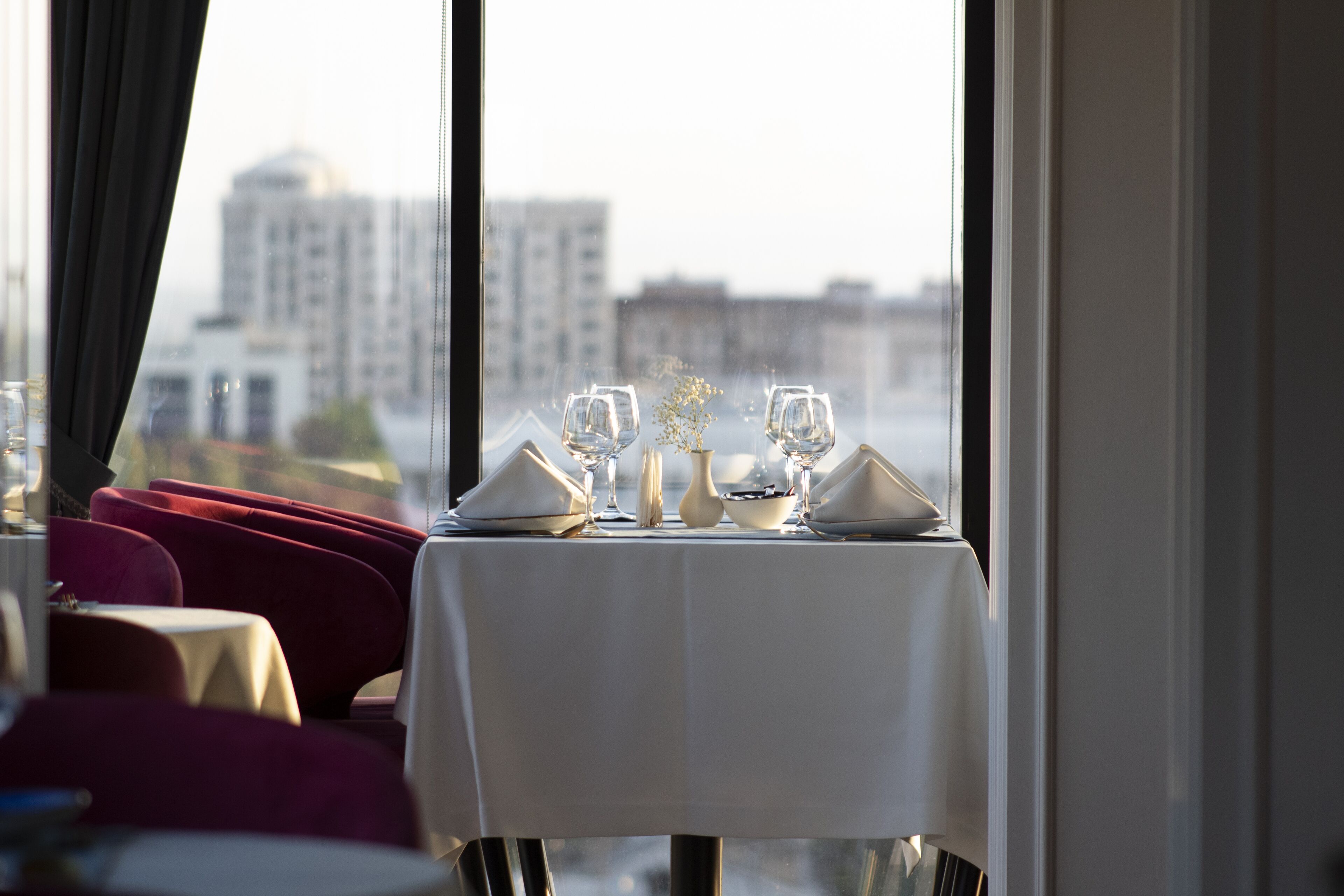 A dining area with a table set for two, a vase with flowers, and a cityscape view through large windows.