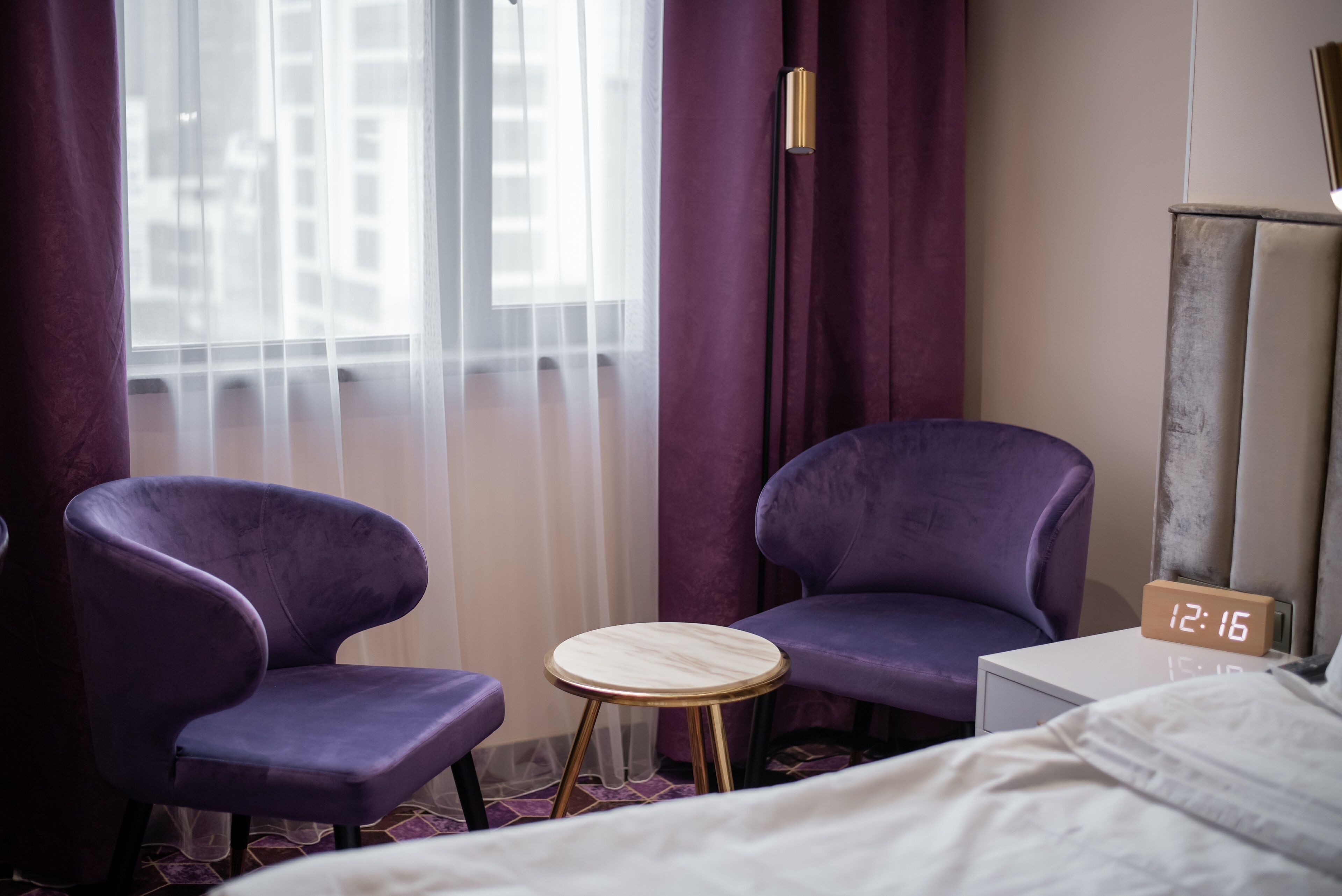 A hotel room with two purple armchairs, a small wooden side table, a bed, a nightstand with a clock, and a window with sheer curtains.