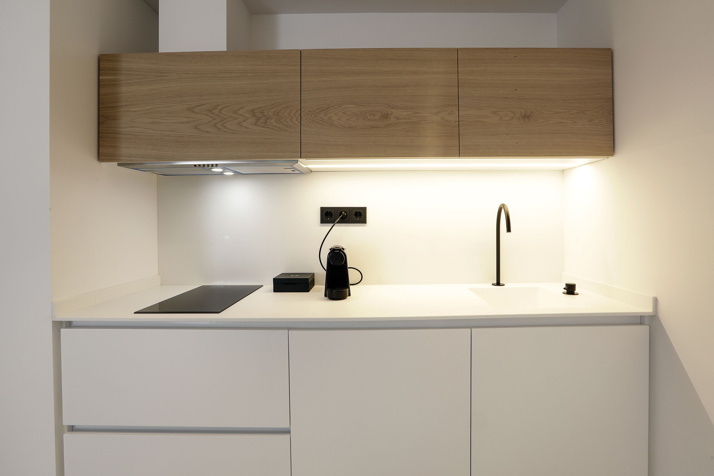 Una cocina moderna con armarios de madera, encimera blanca, cafetera negra y campana extractora integrada.