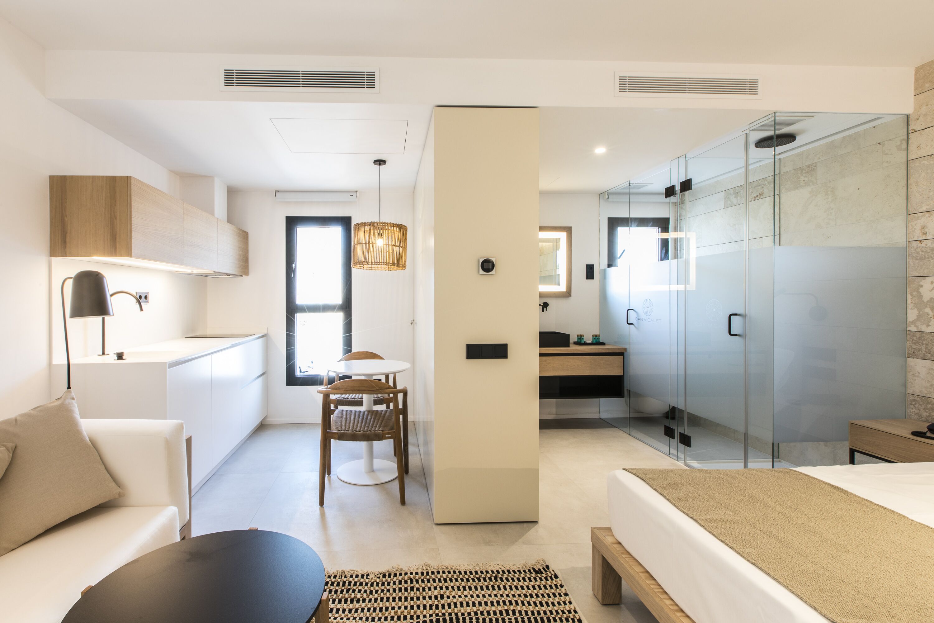 Una habitación de hotel moderna con kitchenette, comedor, dormitorio con cama grande y zona de ducha con paredes de vidrio.