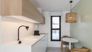 Une cuisine moderne avec un plan de travail blanc, un robinet noir et des armoires en bois. Une fenêtre offre une vue sur les arbres, et un luminaire suspendu en osier complète l’ensemble.