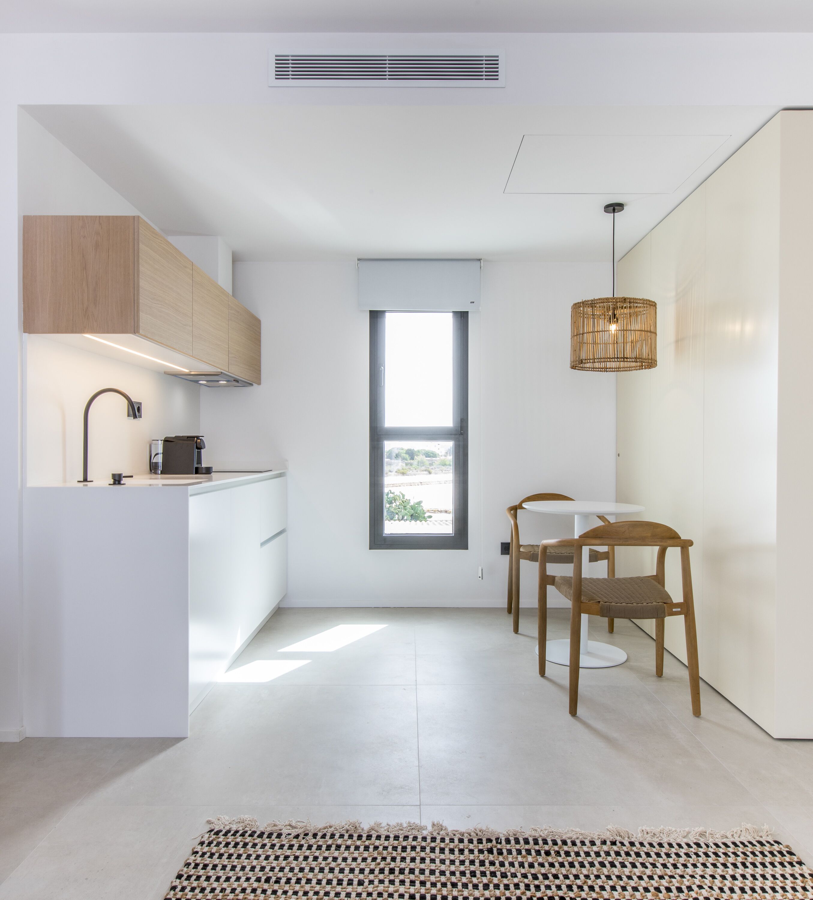 Una cocina moderna con armarios blancos, estufa integrada y una ventana. Hay dos sillas de madera y una mesa pequeña.