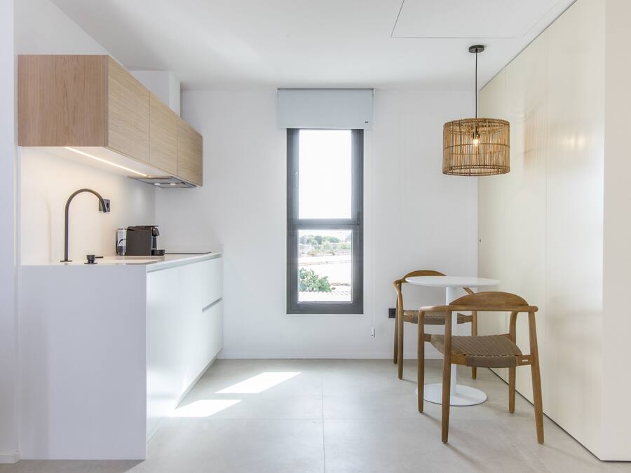 Una cocina moderna con armarios blancos, estufa integrada y una ventana. Hay dos sillas de madera y una mesa pequeña.