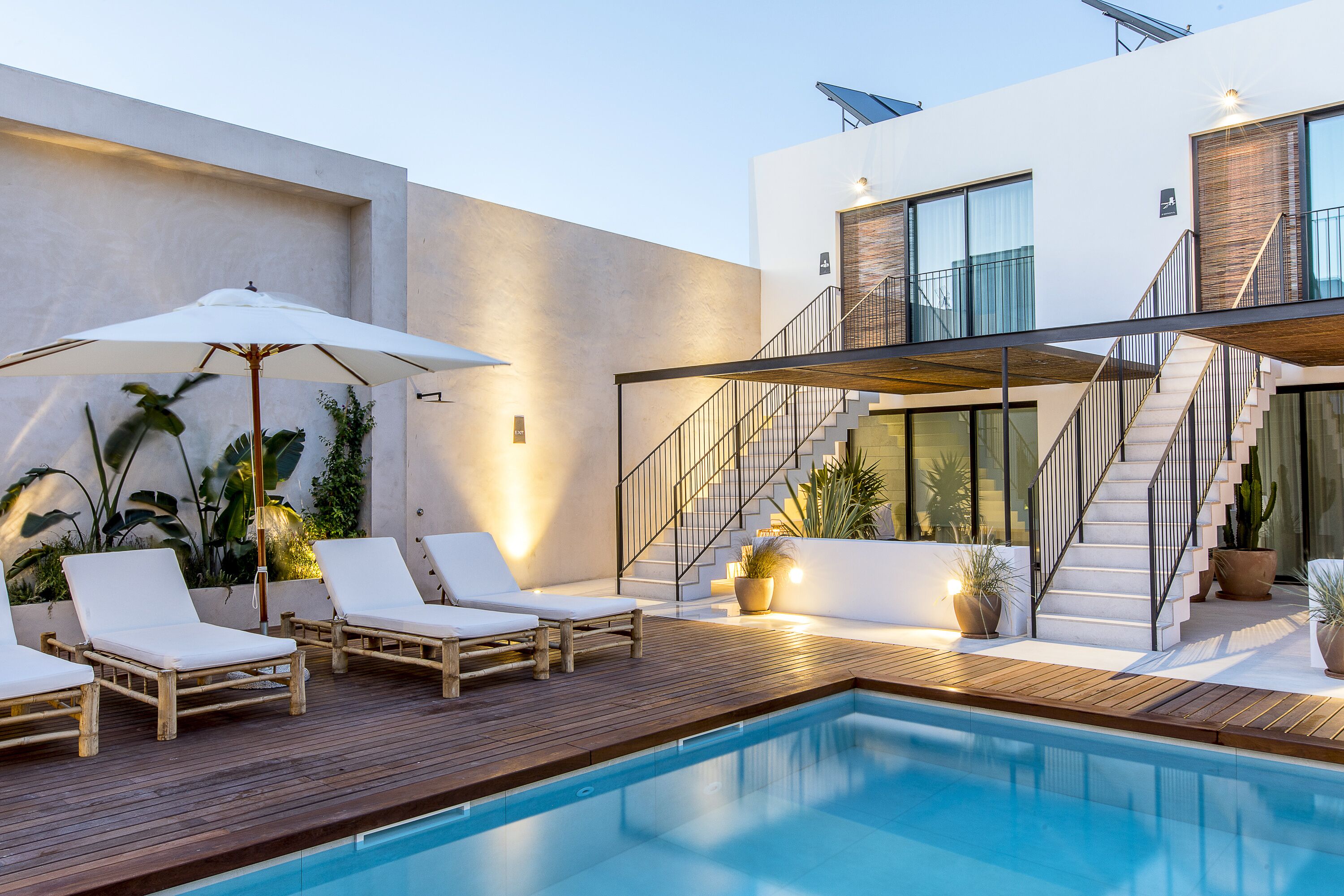 Un área de piscina moderna al aire libre con sillones de descanso y sombrillas, una terraza de madera y una escalera que conduce a un segundo piso.