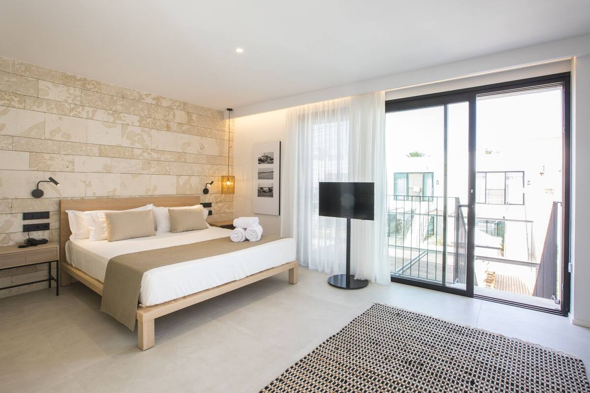 A modern hotel room with a large bed, a bedside table, a floor lamp, and a view of the outside balcony.