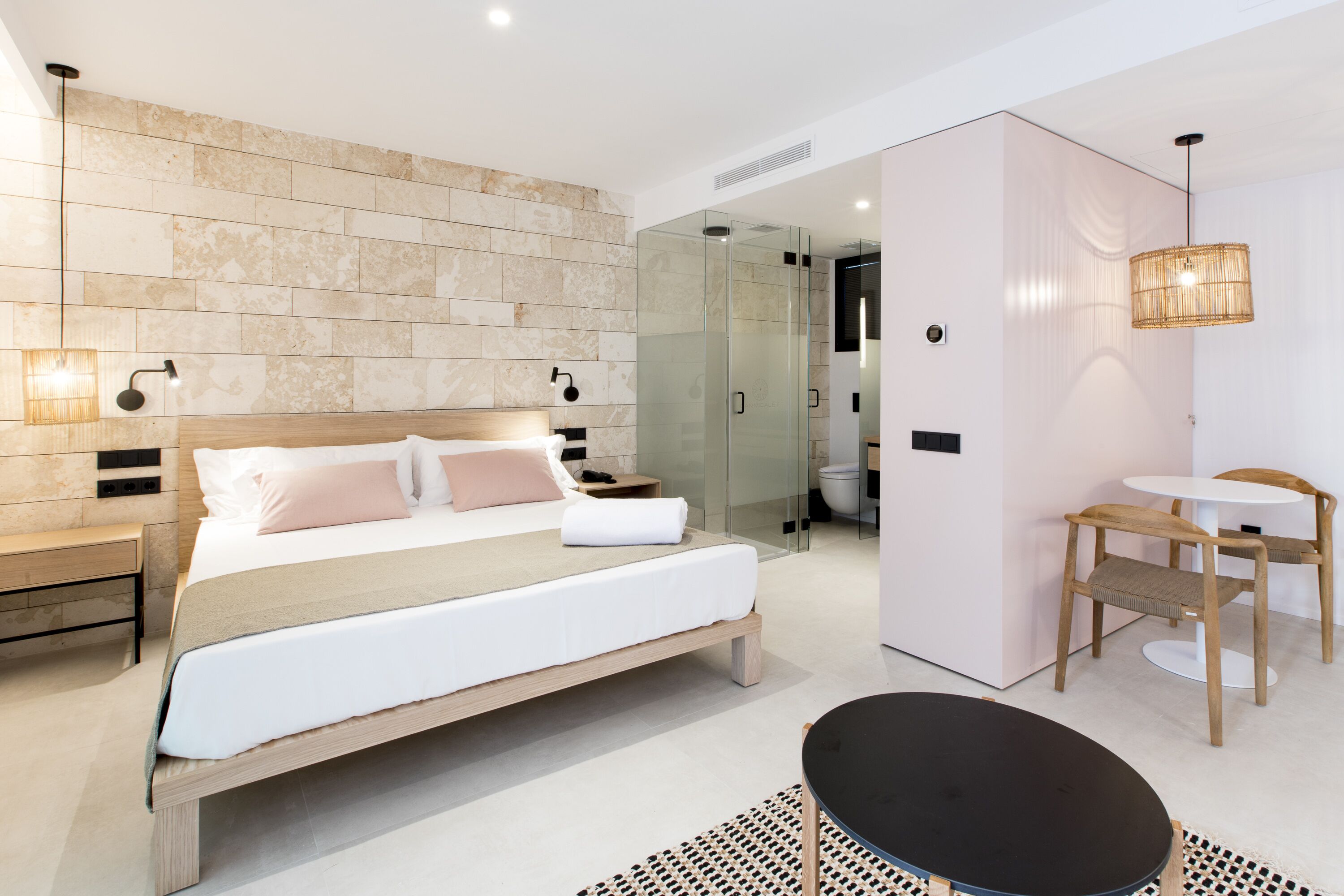 Un dormitorio moderno con una cama grande, mesitas de noche, una ducha con mampara de vidrio, una pequeña mesa redonda y una silla.
