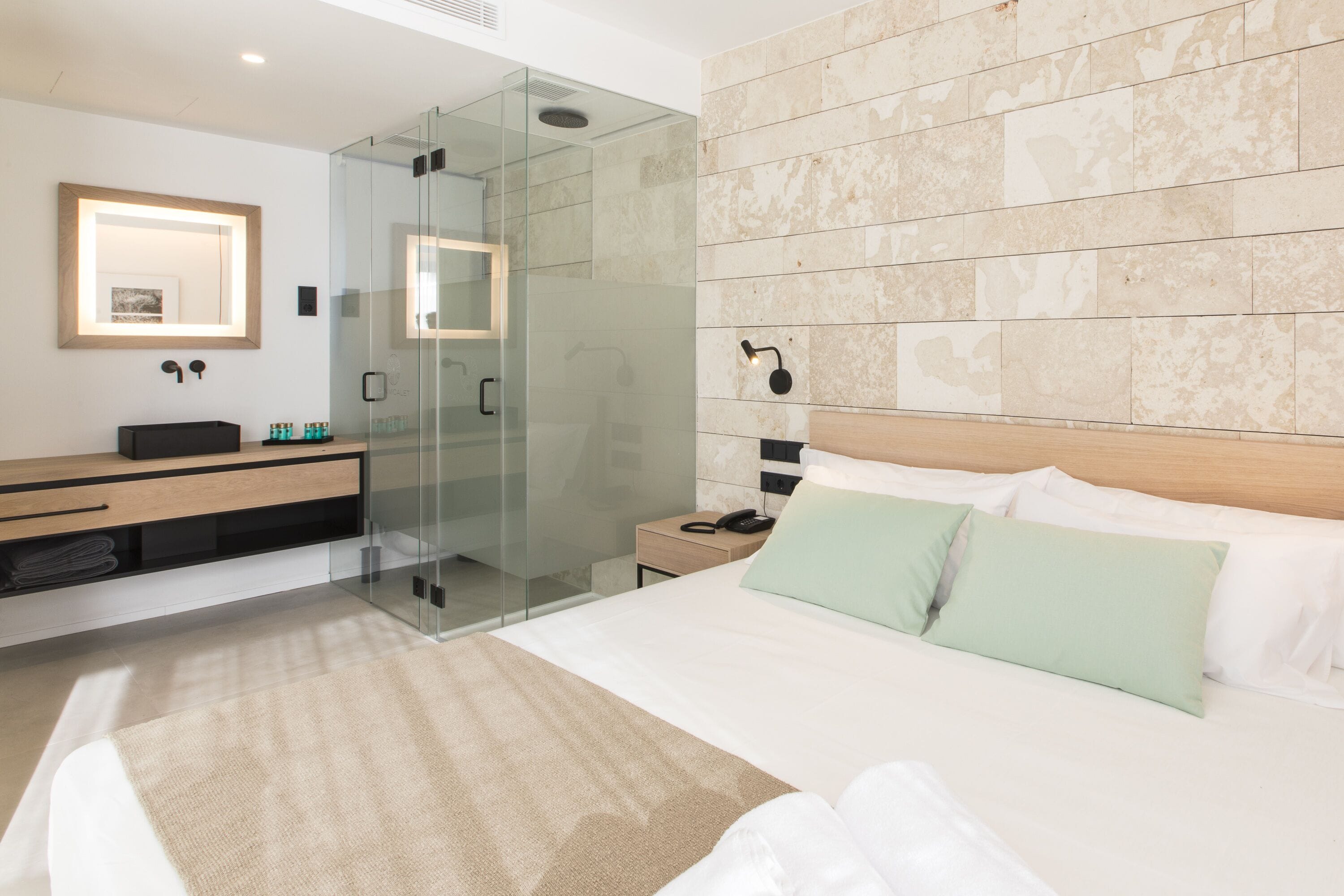 A modern hotel room with a glass-enclosed shower, a wooden headboard, and a large bed with green pillows.