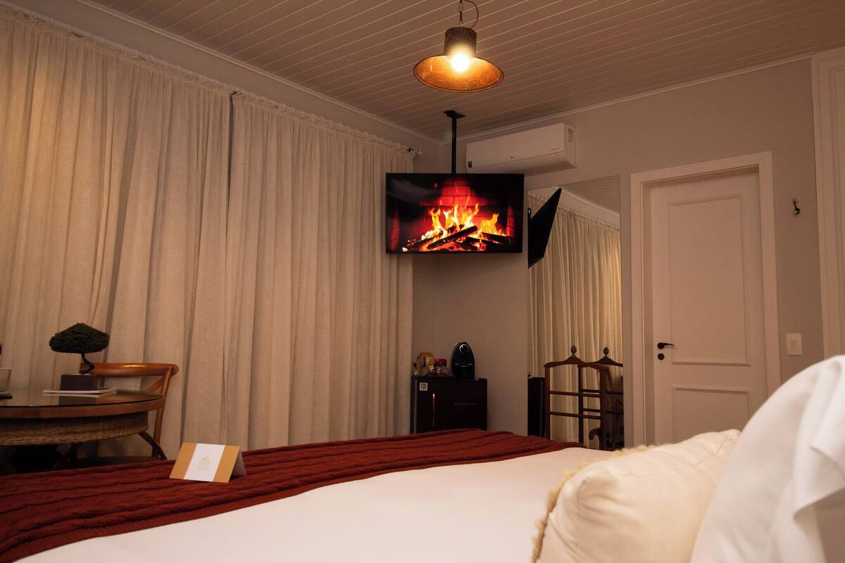 A hotel room with a bed, a flat-screen TV mounted on the wall, a small round table with a chair, a bedside table with a lamp, and a door.