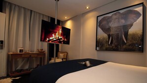 Una habitación de hotel con una cama, un televisor mostrando un fuego, un cuadro de un elefante enmarcado, un escritorio con una lámpara y una silla.