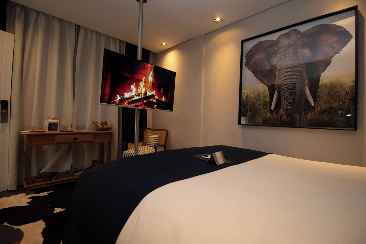 A hotel room with a bed, a television displaying a fire, a framed elephant picture, a desk with a lamp, and a chair.