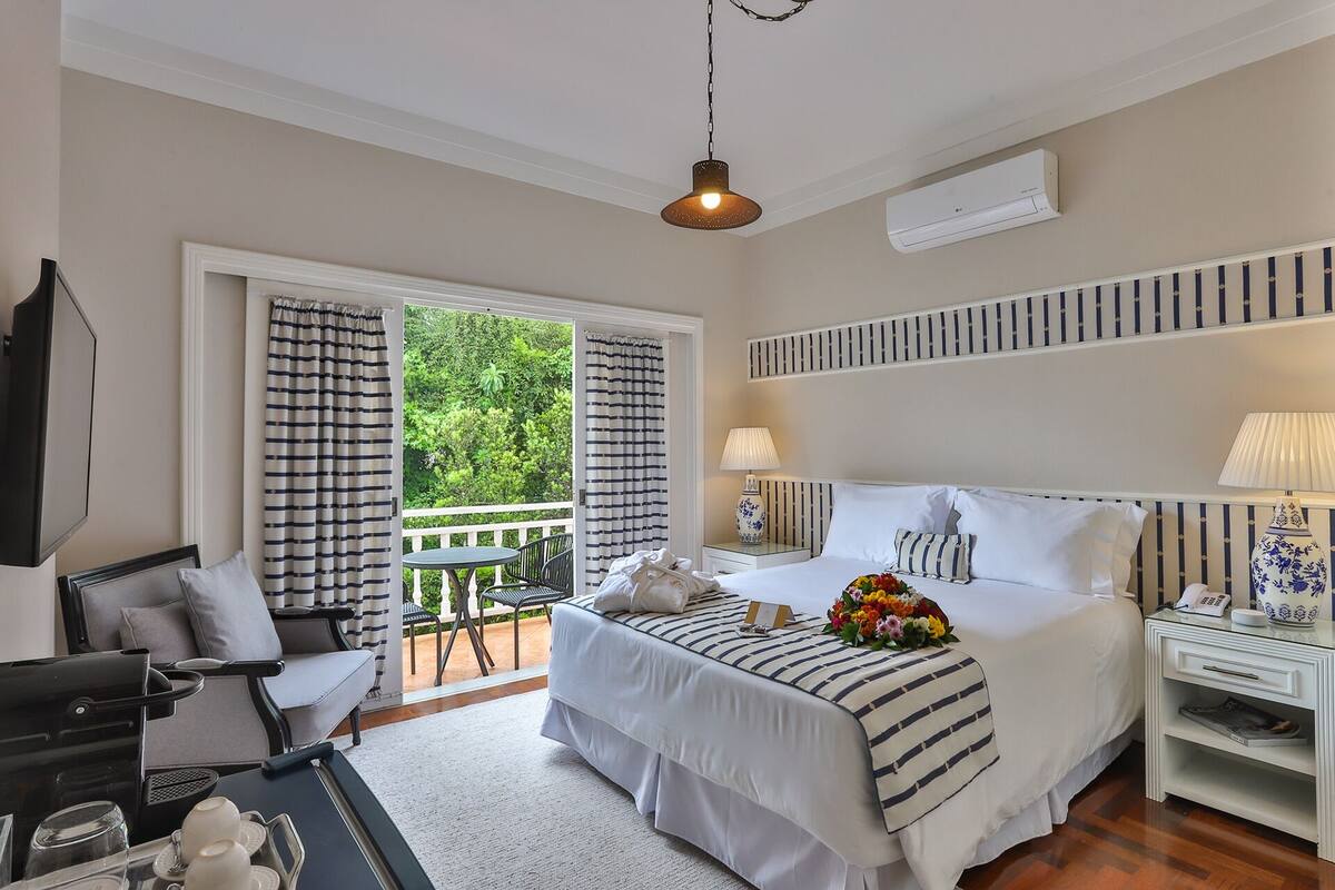 A hotel room with a bed, a desk, a chair, a table with a tray, a hanging light fixture, and a view of greenery through the door.