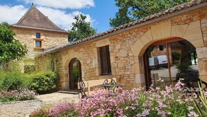 A stone building with arched windows and a tiled roof, surrounded by a garden with pink flowers. - Domaine du Cardou (Puy-l'Eveque)