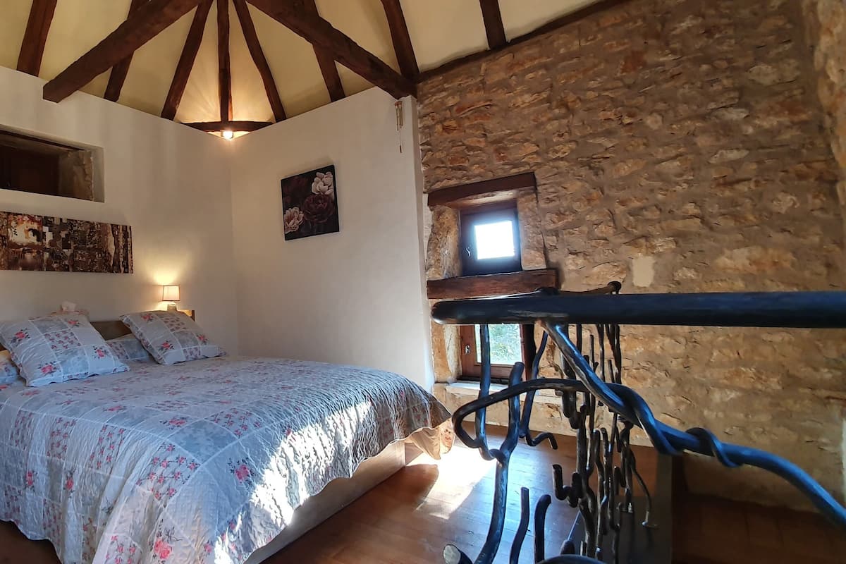 Une chambre avec un lit, des poutres en bois, un mur en pierre et une petite fenêtre.