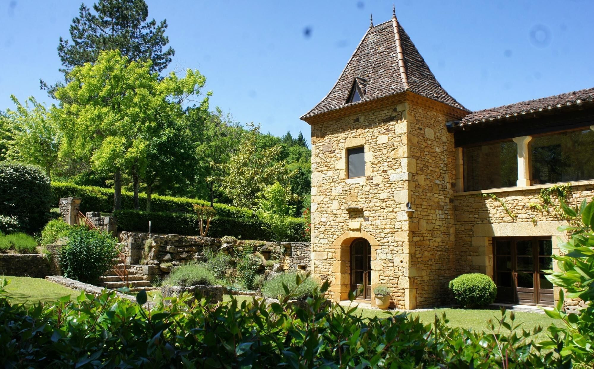 A stone tower with a pitched roof, a stone building with large windows, and a well-maintained garden with a variety of plants and a stone wall.