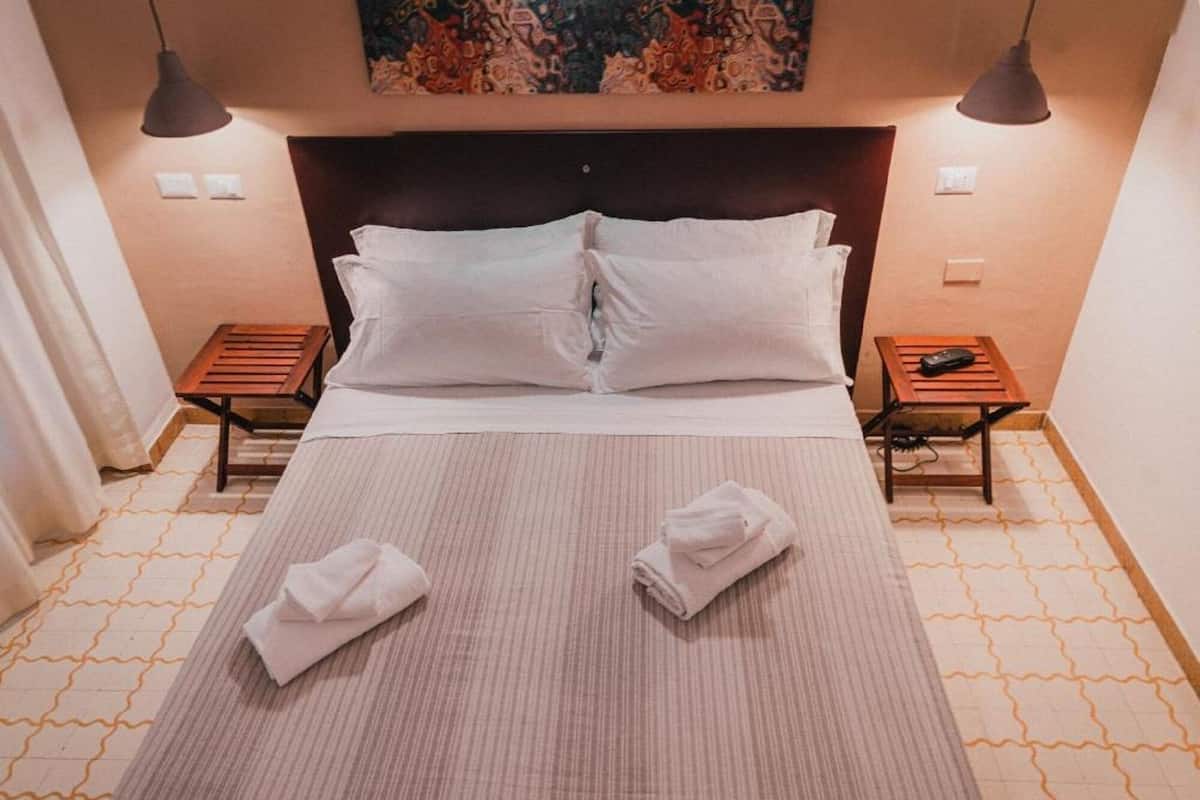 A neatly made bed with white linens and two towels folded on it, two bedside tables with lamps, and a painting above the bed.