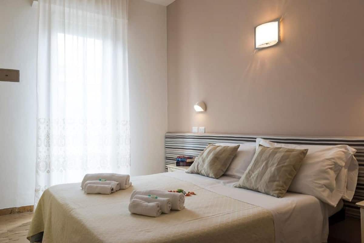 A hotel room with a large bed, two pillows, a bedside table, a wall-mounted light, and a window with sheer curtains.