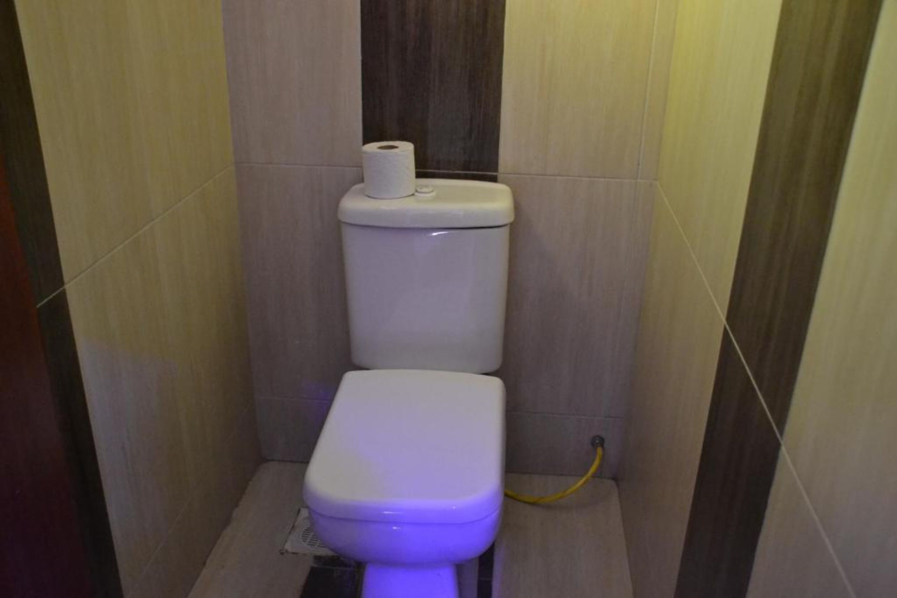 A small, modern bathroom with a toilet, a roll of toilet paper on the tank, and a yellow hose connected to the wall.