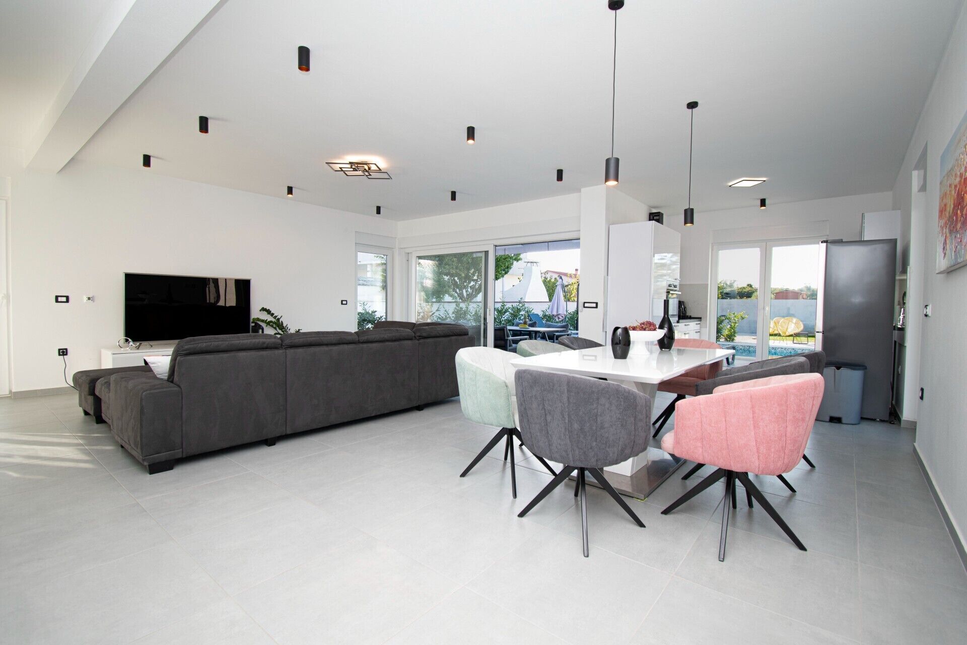 Un salon moderne avec un grand canapé d’angle gris, un coin repas comprenant une table blanche et quatre chaises, un téléviseur à écran plat et une cuisine équipée d’un réfrigérateur.