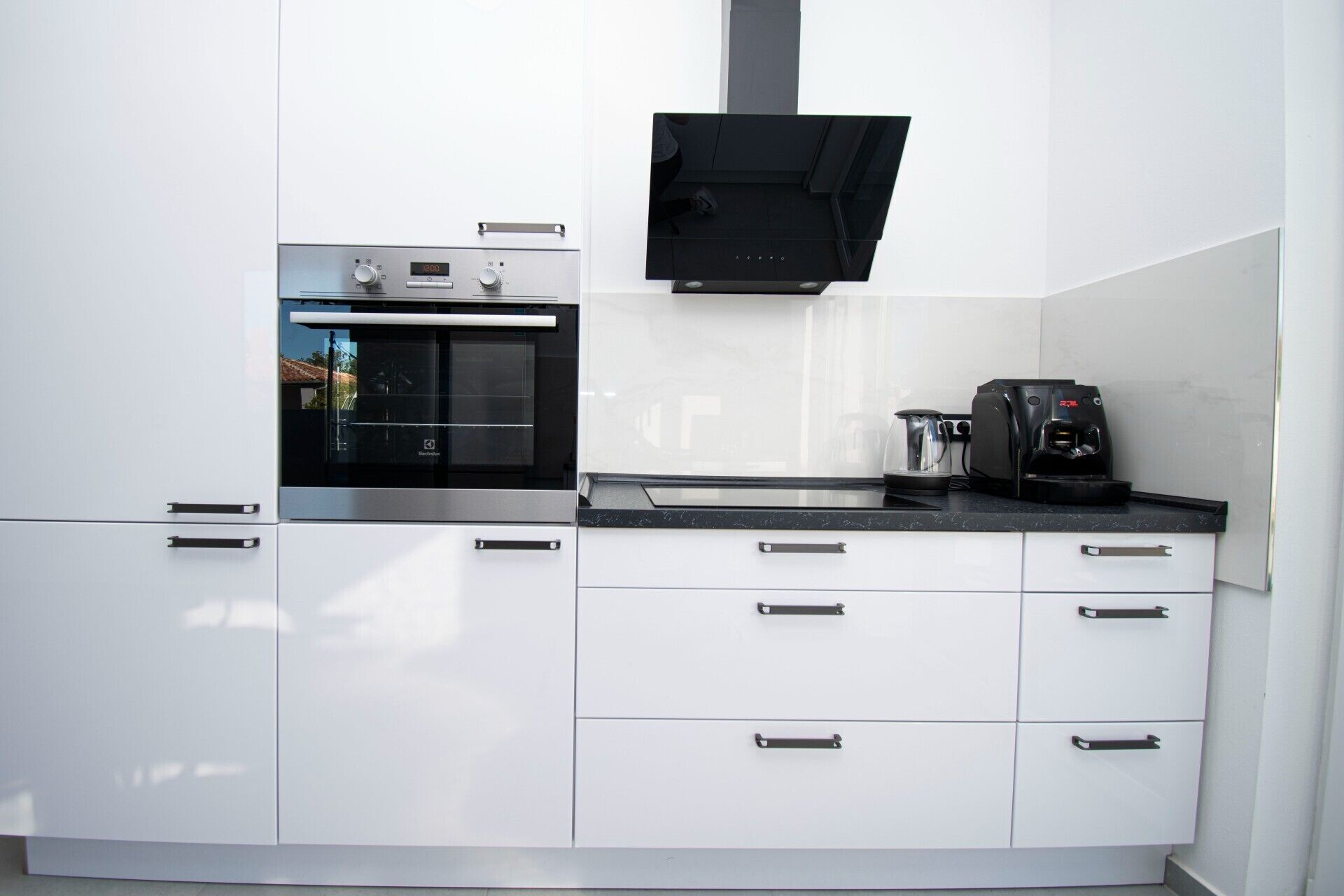 Eine moderne Küche mit weißen Schränken, einem eingebauten Backofen, einer Kaffeemaschine und einer schwarzen Dunstabzugshaube.