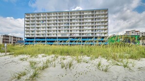 A multi-story hotel with a flat roof and balconies, located near a sandy beach with tall grass. - Coastal Condo w/ Balcony in North Myrtle Beach! (North Myrtle Beach)