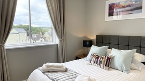Un lit bien fait, avec une tête de lit matelassée, une lampe de chevet et une vue sur l’extérieur par une fenêtre ornée de rideaux.