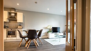 Une cuisine moderne dotée d’appareils électroménagers intégrés, un coin repas avec une table et des chaises en verre, et un espace de vie agrémenté d’un téléviseur et de plantes décoratives.