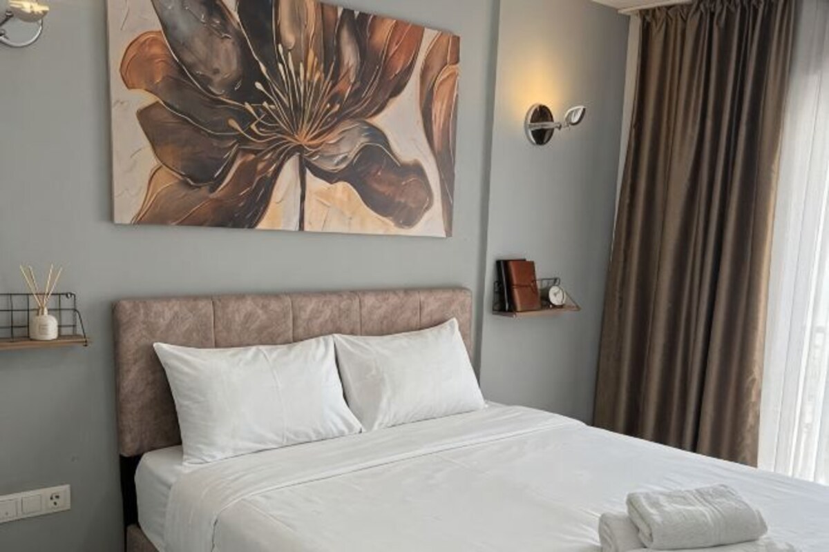 A neatly made bed with white linens, a large floral artwork on the wall, a bedside table with a lamp, and a window with curtains.