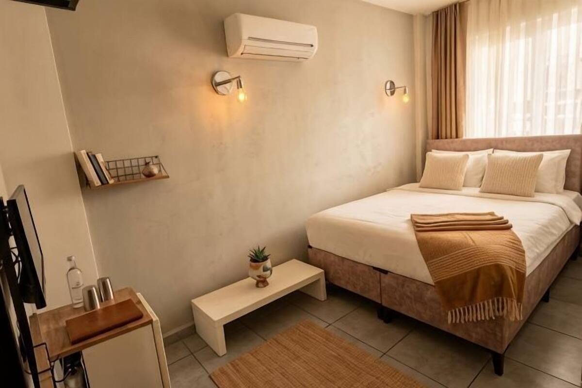 Un dormitorio con cama, mesita de noche, banco, aire acondicionado instalado en la pared y un estante pequeño con libros y una planta.