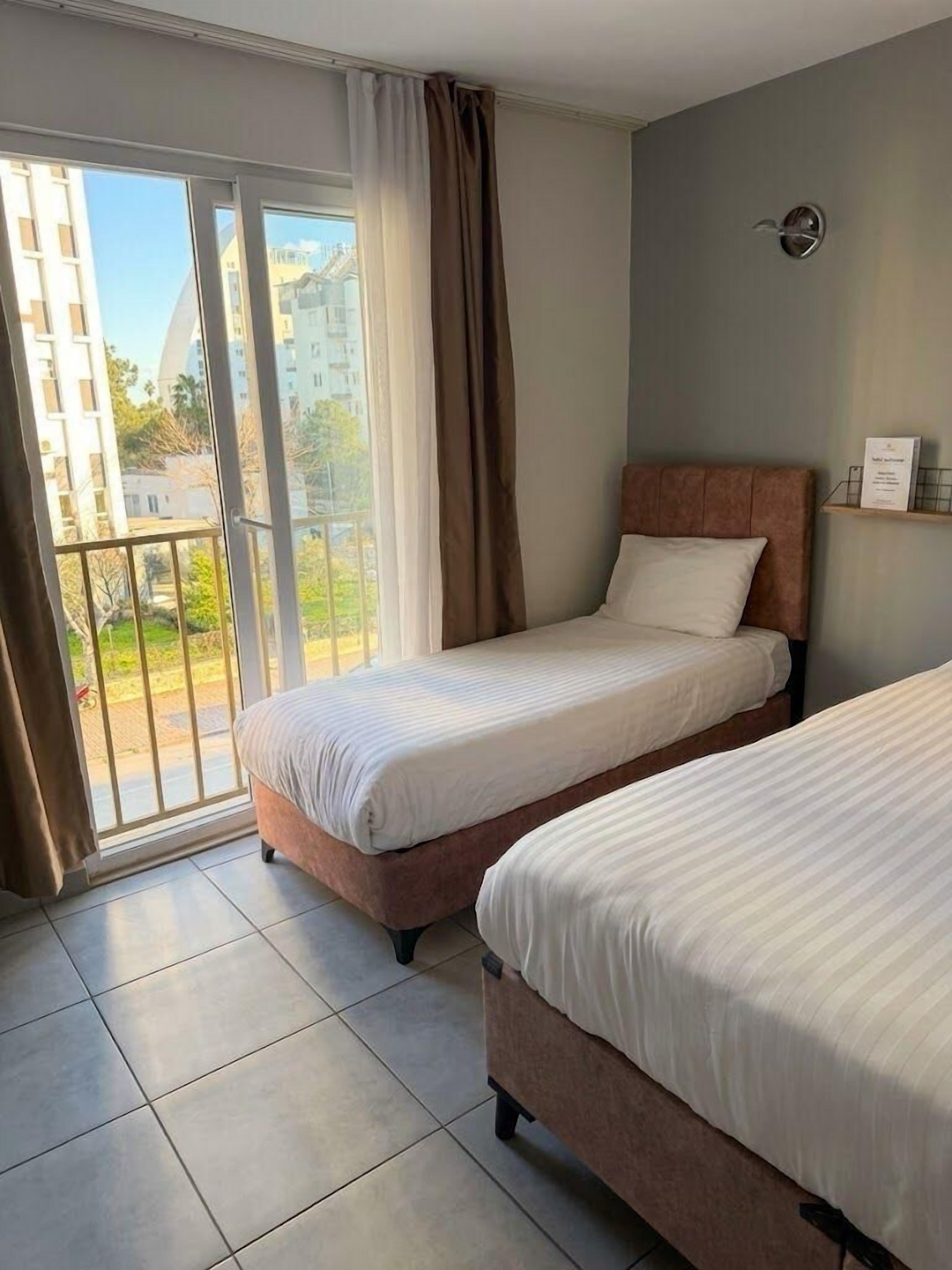 A hotel room with two beds, a sliding glass door to the outside, and a view of buildings and greenery.