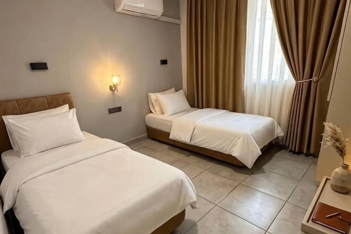 Habitación de hotel con dos camas, una lámpara de pared, una ventana con cortinas y una pequeña mesa con un jarrón.