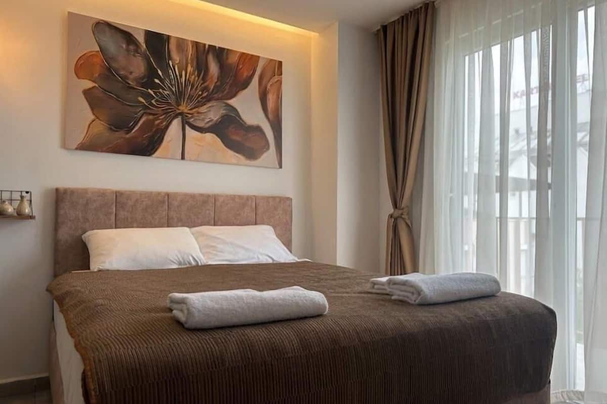 Una habitación de hotel con una cama grande, cabecera, un cuadro de una flor y una ventana con cortinas.