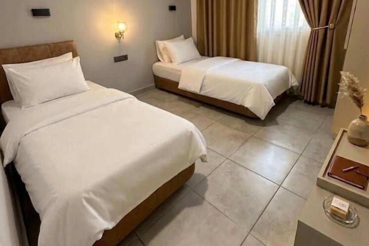 Habitación de hotel con dos camas, aire acondicionado de pared, lámpara de noche y ventana con cortinas.
