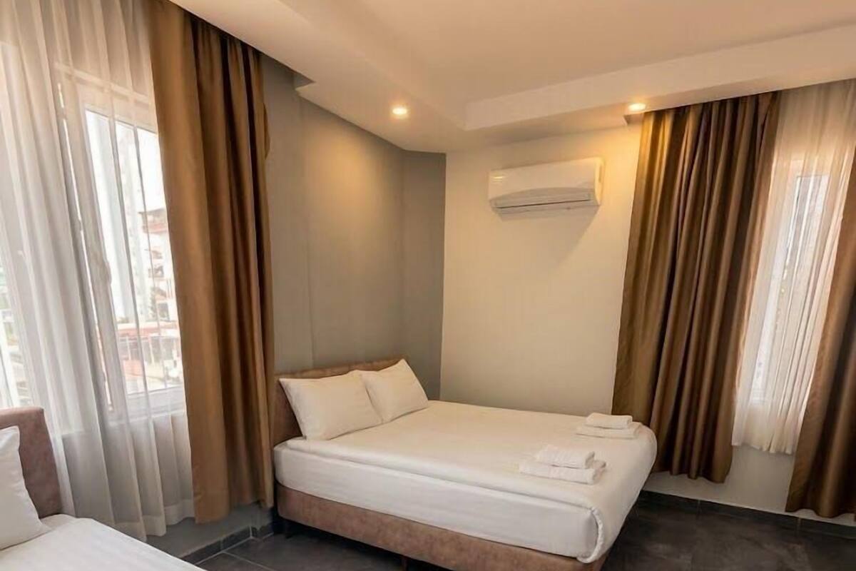 Habitación de hotel con dos camas, un ventanal con cortinas, un equipo de aire acondicionado y piso de baldosas.