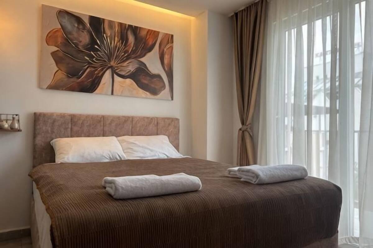 Una habitación de hotel con una cama grande, un cabecero, un cuadro en la pared y una ventana con cortinas.