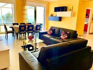 A living room with a sofa, coffee table, and dining area. There is a sliding glass door leading to a balcony.