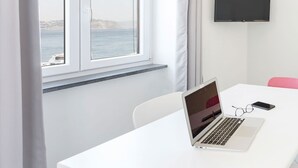 A room with a white table, a laptop, and a smartphone on it. There is a window with curtains and a view of the sea. A flat-screen TV is mounted on the wall. - Marina Grande - Gioia Apartments (Procida)