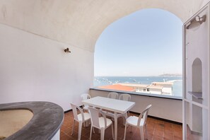 Een balkon met een witte tafel en stoelen, een kleine verwarming en uitzicht op zee met boten.